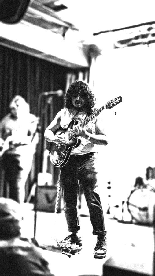A high-contrast black and white photograph of a guitarist playing a hollow-body electric guitar, shot from the crowd.
