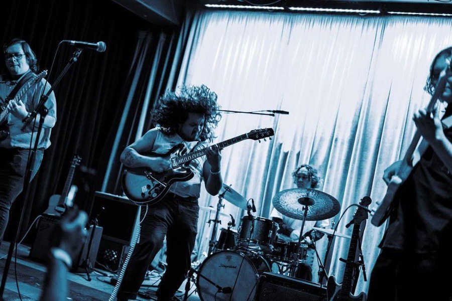 A blue-tinted photograph of the band onstage in front of a bright curtain, shot from among the audience.